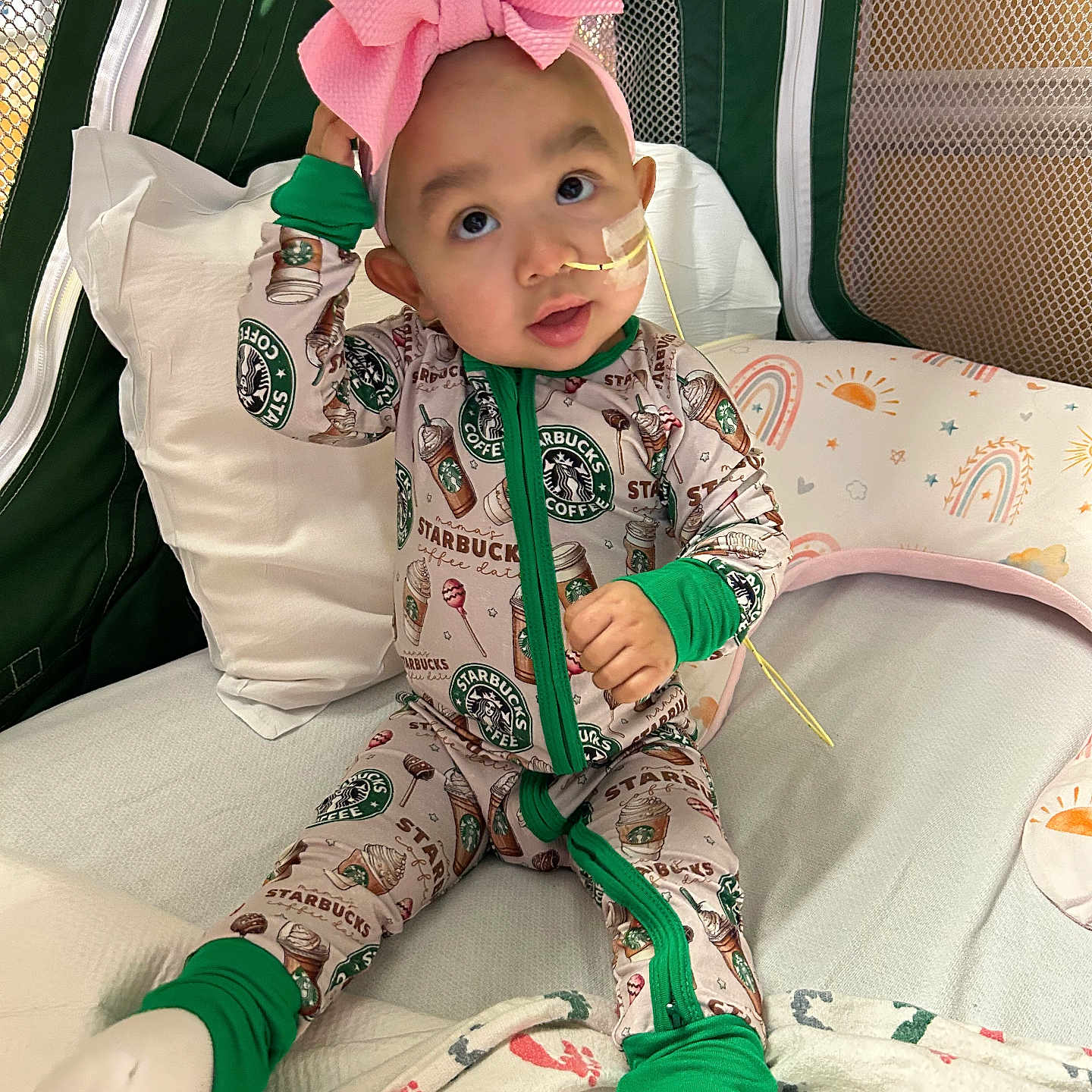 Anahi is registered to the contest to win money with this photo: baby, child, onesie, headband, pink_bow, starbucks_logo, feeding_tube, bed, pillow, blanket, footwear, green_trim, cute, indoor, infant, sitting, expression, curious, person, cozy