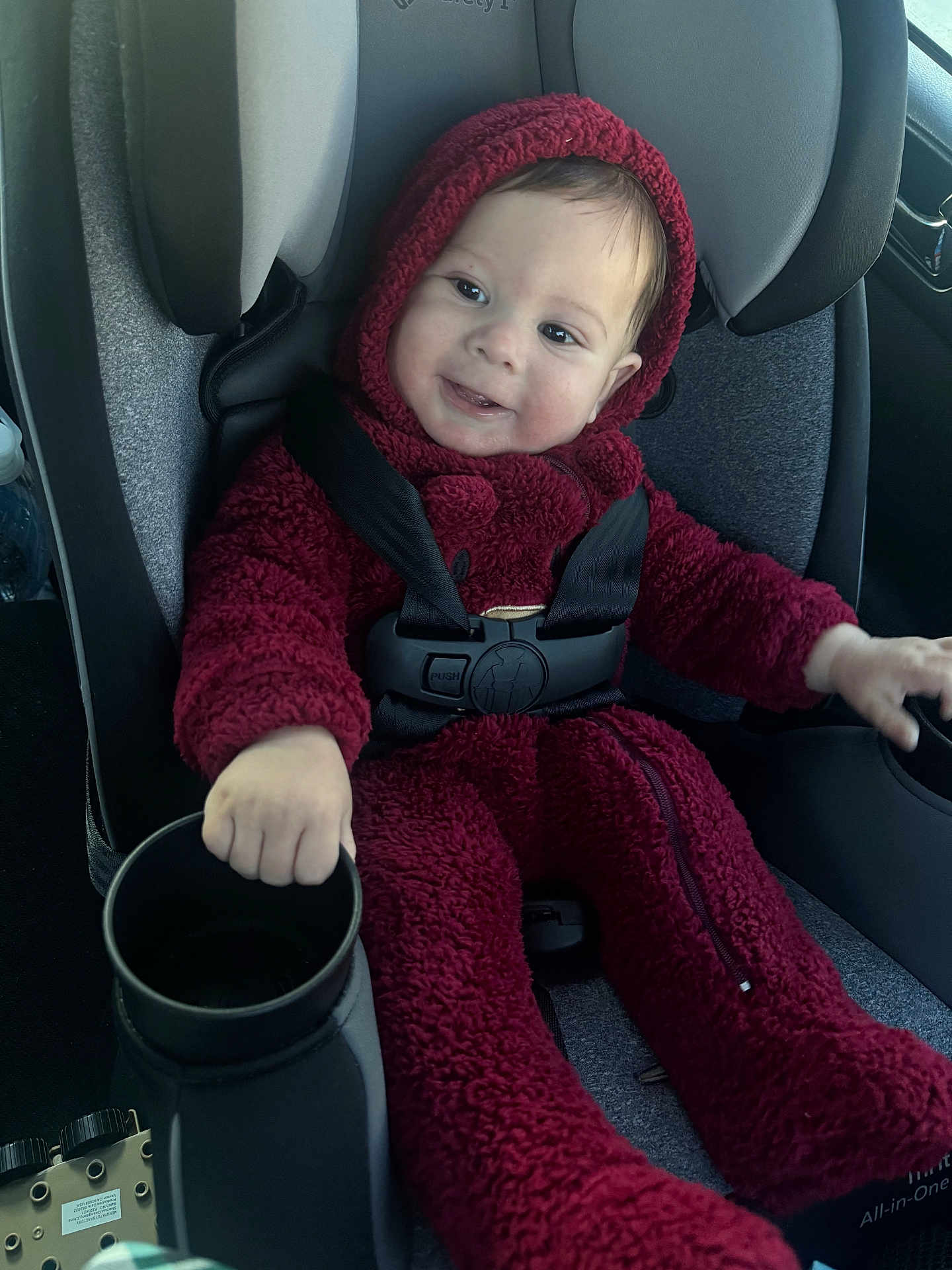 Enzo is registered to the contest to win money with this photo: baby, child, car_seat, red_onesie, hoodie, smiling, seatbelt, cup_holder, car_interior, safety, infant, happy, portrait, cozy, clothing, person, baby_fashion, seated, cute, transportation