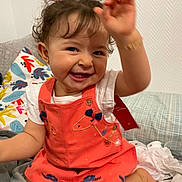 Lydia participe au concours pour gagner de l'argent avec cette photo : toddler, child, smiling, happy, curly_hair, pigtails, dress, coral_dress, embroidered, hand_up, indoor, bed, pillows, blanket, white_shirt, cute, playful, person, portrait, young_child