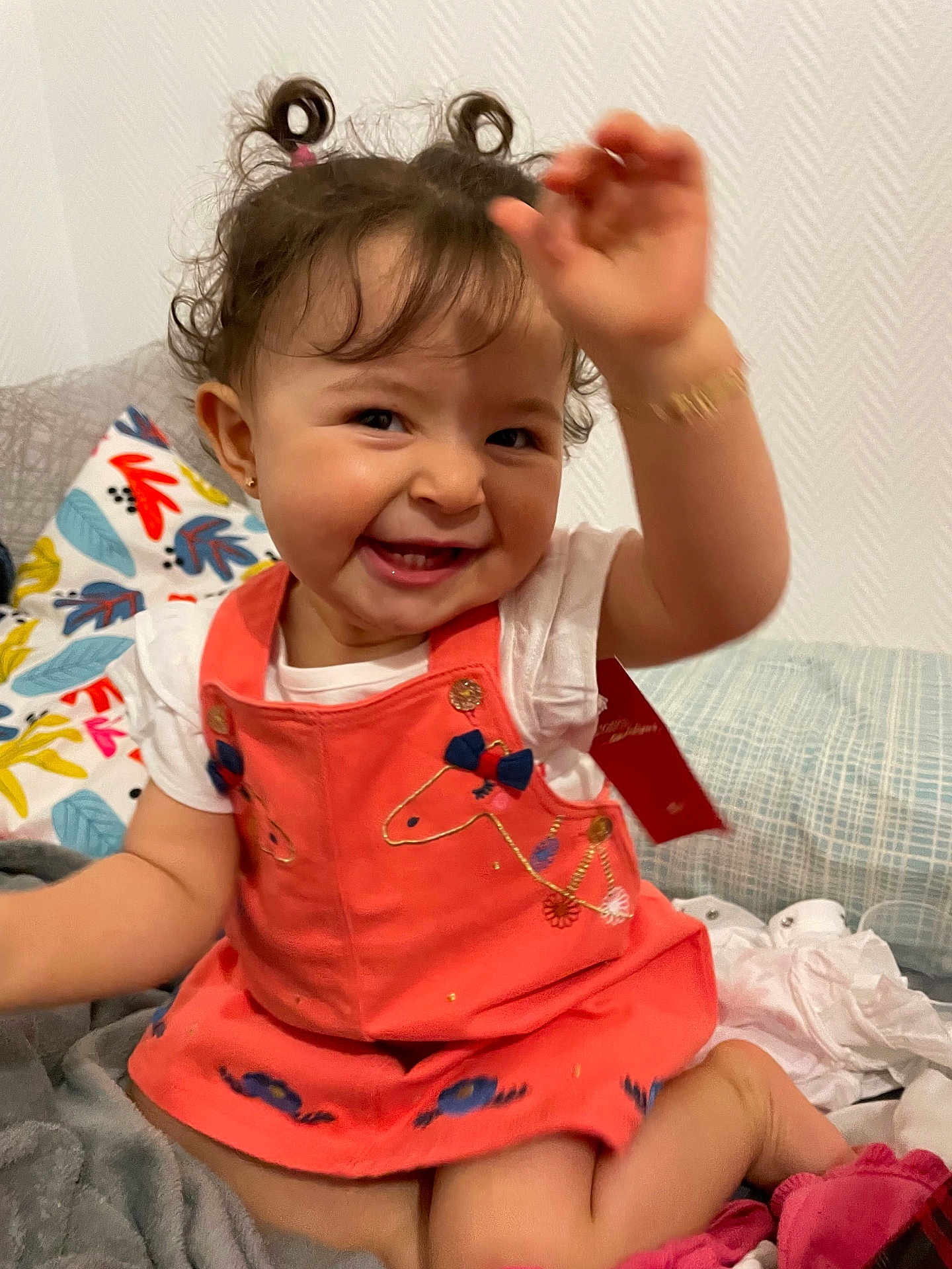 Lydia participe au concours pour gagner de l'argent avec cette photo : toddler, child, smiling, happy, curly_hair, pigtails, dress, coral_dress, embroidered, hand_up, indoor, bed, pillows, blanket, white_shirt, cute, playful, person, portrait, young_child