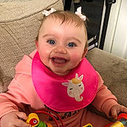 Skyla joined the competition — help win amazing prizes! baby, bib, blue_eyes, bow, child, closeup, colorful_toy, couch, elephant_unicorn, hair_bows, hands, happy, indoor, pink_bib, pink_clothing, portrait, smiling, toddler, toy, upholstery