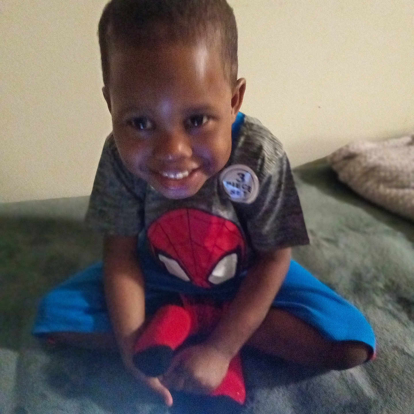 Steve Williams is registered to the contest to win money with this photo: child, smiling, spiderman, plush_toy, bed, blanket, indoor, casual_clothing, person, happy, sitting, young, cute, toy, portrait, home, comfortable, brown_skin, short_hair, tshirt
