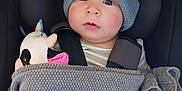 Kenny is registered to the contest to win money with this photo: baby, child, hat, knit_hat, pom_pom, blanket, car_seat, plush_toy, stuffed_animal, cow_toy, clothing, face, headwear, bundle, warm, infant, seatbelt, closeup, indoor, portrait