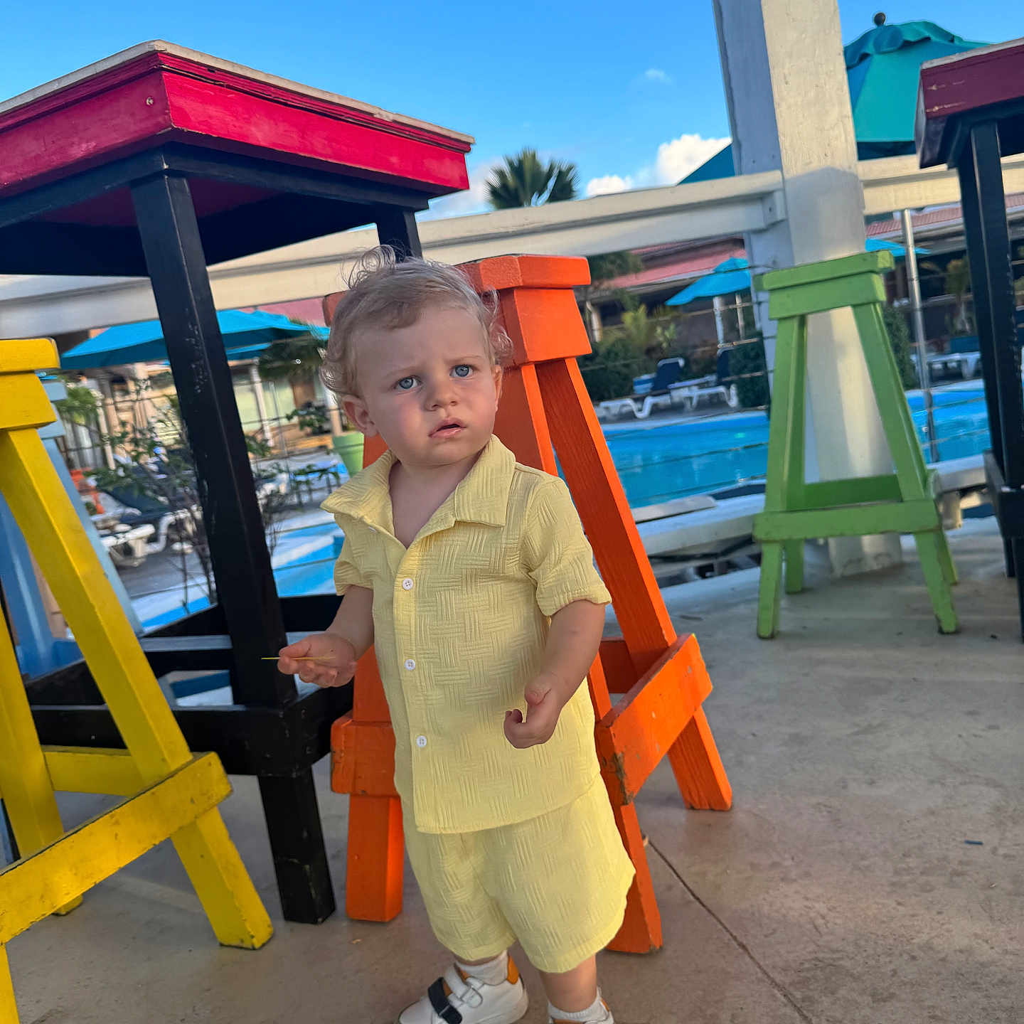 Lyor a rejoint le concours — aidez-le/la à gagner de superbes lots ! boy, button_shirt, child, colorful_stools, curious_expression, curly_hair, orange_stool, outdoor, pavement, pool, portrait, shorts, sneakers, standing, sunny, toddler, umbrella, vacation, yellow_outfit, yellow_stool