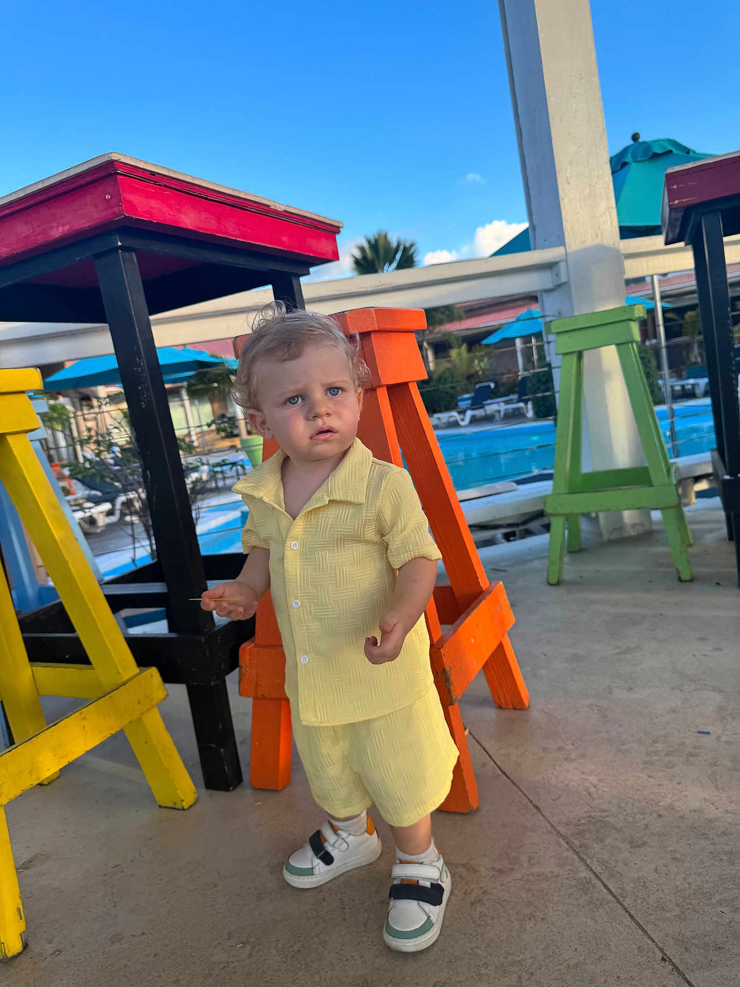 Lyor a rejoint le concours — aidez-le/la à gagner de superbes lots ! child, toddler, boy, yellow_outfit, shorts, button_shirt, sneakers, curly_hair, colorful_stools, orange_stool, yellow_stool, pool, umbrella, outdoor, sunny, pavement, portrait, curious_expression, vacation, standing