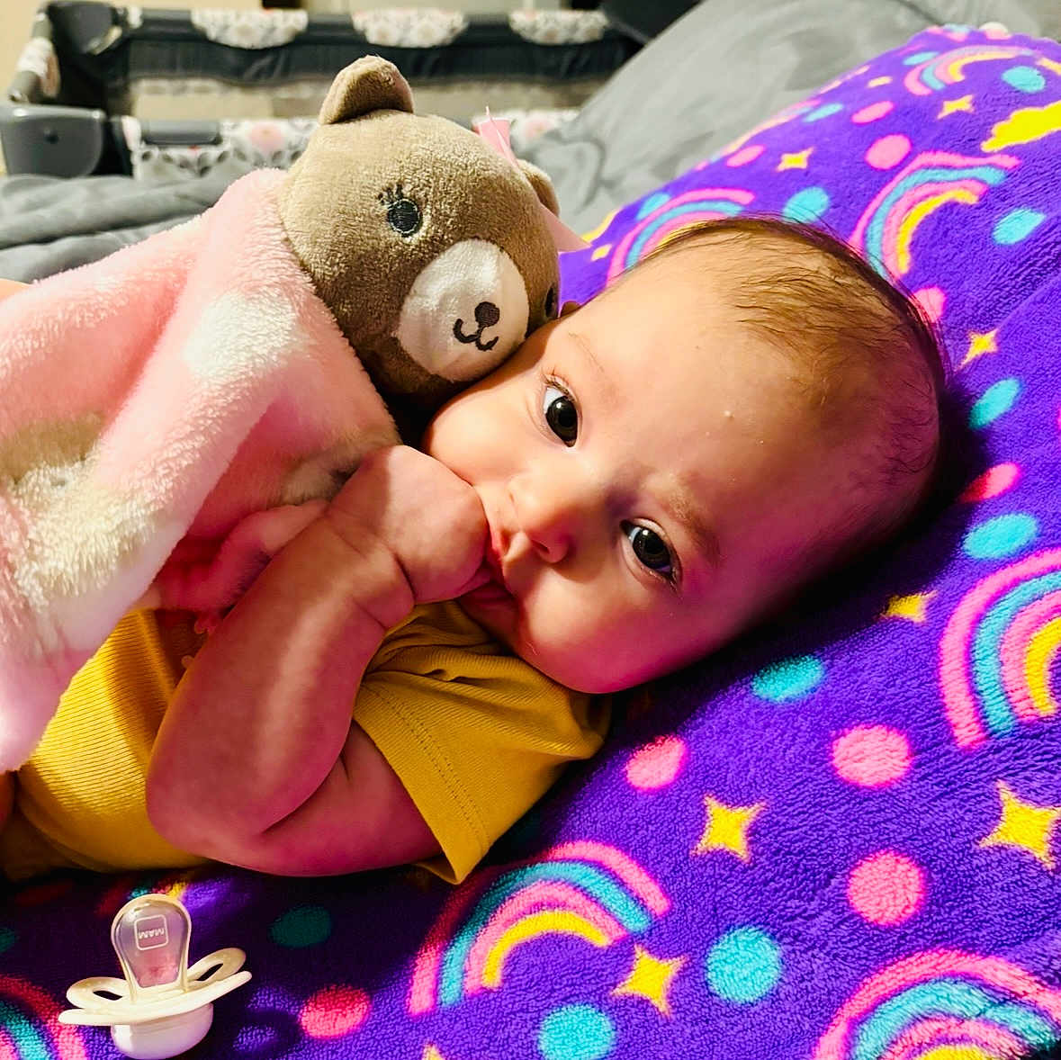 Addison is registered to the contest to win money with this photo: baby, infant, teddy_bear, blanket, purple_blanket, rainbow_pattern, stars, thumb_sucking, pacifier, yellow_clothing, cute, cozy, soft_toy, lying_down, indoor, child, face, head, sleepy, comfort