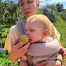 baby, parent, child, infant, apple, fruit, front_carrier, outdoor, garden, nature, sunlight, blue_sky, greenery, portrait, family, holding, eating, blond_hair, eyeglasses, smiling