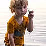 boy, casual_clothing, child, curious, daylight, nature, orange_shirt, outdoor, person, play, portrait, reflection, ripples, small_object, standing, summer, water, water_drip, wet_hair, young