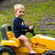 Blake is registered to the contest to win money with this photo: child, blonde_hair, barefoot, toy_vehicle, yellow, outdoor, grass, gravel, long_sleeve_shirt, sitting, vehicle, play, kid, casual, daylight, young, bare_legs, small, foot, person