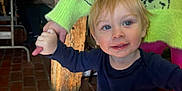 Blake is registered to the contest to win money with this photo: adult_hand, background, blue_eyes, brick_floor, casual_clothing, child, festive_decor, glasses, green_pants, grinch_sweater, holding_hands, indoor, navy_shirt, person, purple_boots, smile, table, toddler, woman, wooden_pillar