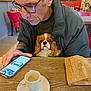 man, dog, cavalier_king_charles_spaniel, glasses, jacket, smartphone, table, coffee_cup, spoon, paper_bag, chair, indoor, restaurant, brown_hair, facial_expression, pet, looking, seated, wooden_table, casual