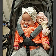 Elyana participe au concours pour gagner de l'argent avec cette photo : baby, pacifier, stroller, headband, bow, orange_jacket, pink_toy, stuffed_animal, smiling, child, infant, clothing, seatbelt, indoor, person, cute, baby_girl, cozy, portrait, toddler
