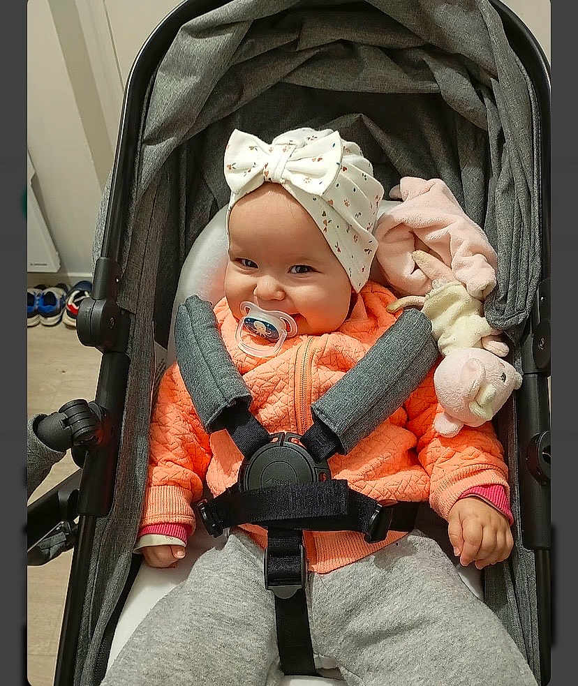 Elyana participe au concours pour gagner de l'argent avec cette photo : baby, pacifier, stroller, headband, bow, orange_jacket, pink_toy, stuffed_animal, smiling, child, infant, clothing, seatbelt, indoor, person, cute, baby_girl, cozy, portrait, toddler