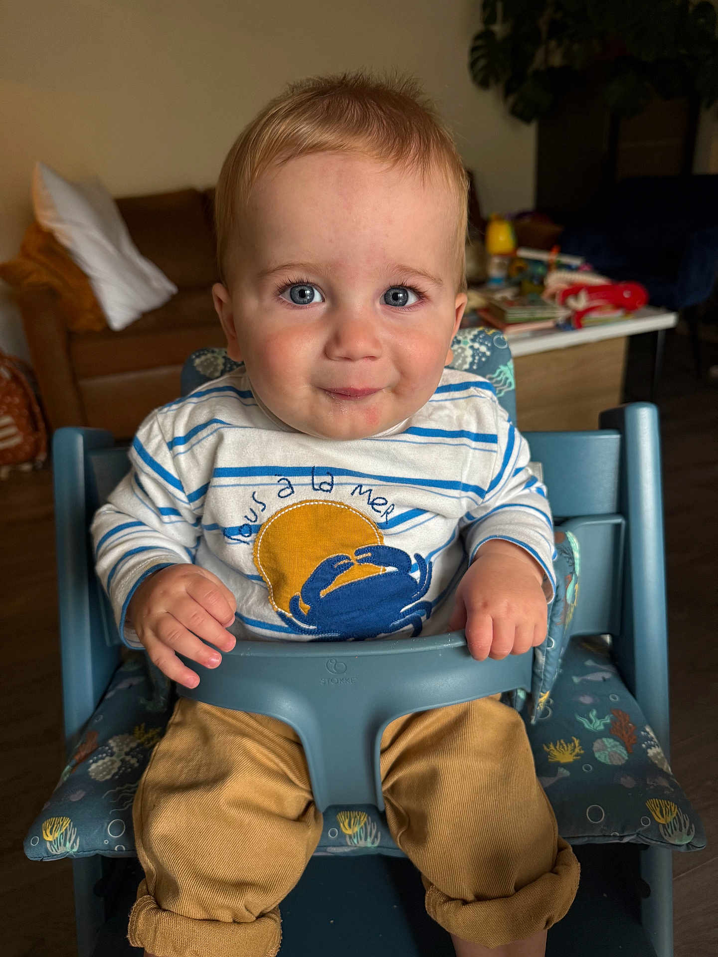 Milo a rejoint le concours — aidez-le/la à gagner de superbes lots ! baby, child, toddler, high_chair, blue_eyes, striped_shirt, crab_design, pants, rolled_up_pants, indoor, couch, cushions, table, toys, person, face, hands, smile, furniture, home