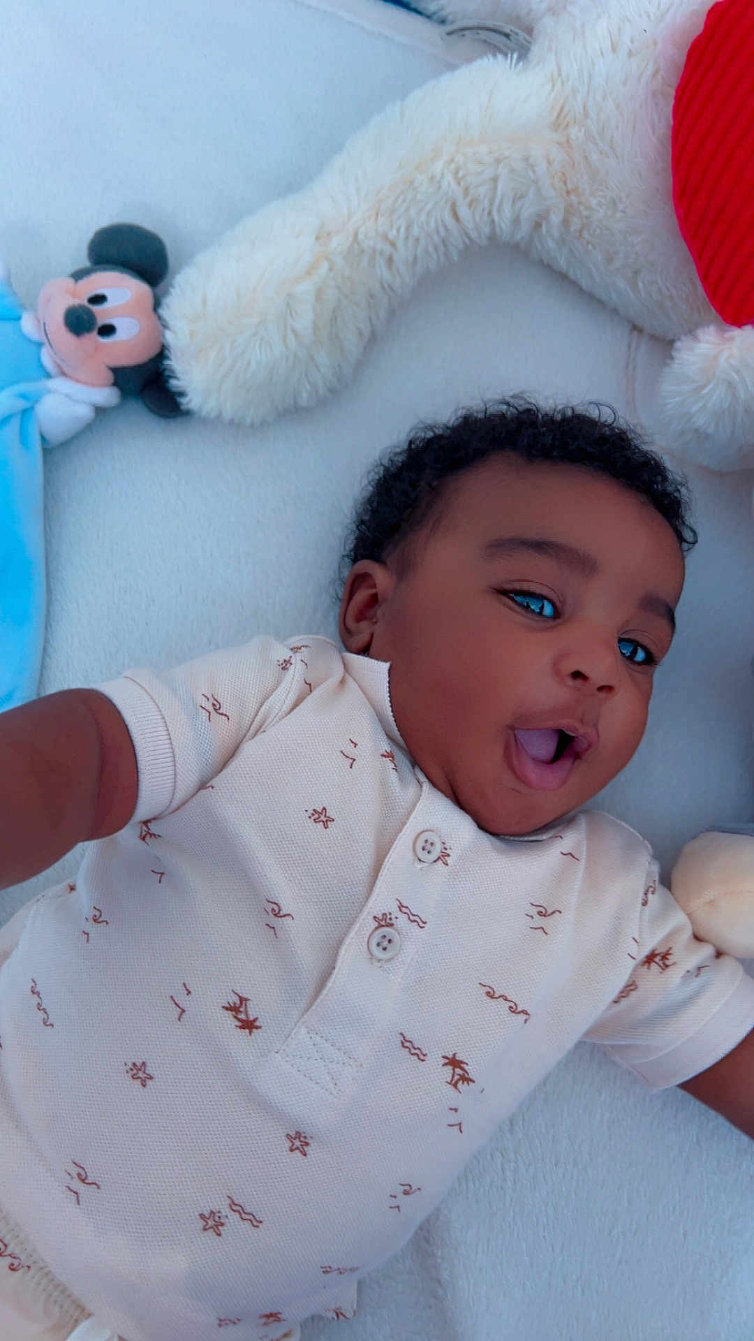 Eli-Nathan a rejoint le concours — aidez-le/la à gagner de superbes lots ! baby, infant, child, smiling, lying_down, plush_toy, mickey_mouse, blanket, soft_texture, cute, happy, indoors, clothing, short_sleeves, curly_hair, portrait, playful, comfort, young_child, resting