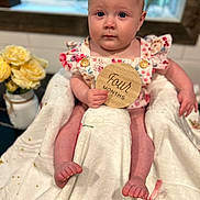 Libby joined the competition — help win amazing prizes! baby, child, infant, blanket, flower, vase, window, wooden_sign, face, person, clothing, sitting, cute, indoor, floral_pattern, skin, hand, foot, table, soft