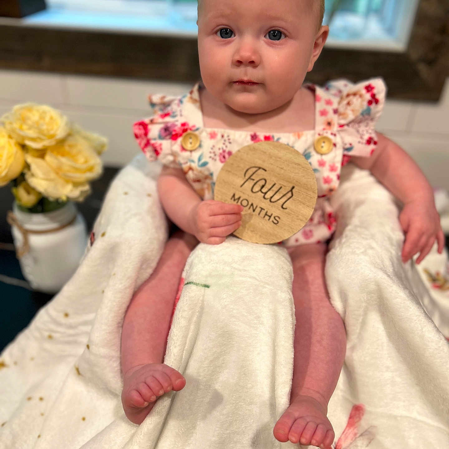 Libby joined the competition — help win amazing prizes! baby, child, infant, blanket, flower, vase, window, wooden_sign, face, person, clothing, sitting, cute, indoor, floral_pattern, skin, hand, foot, table, soft