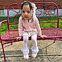 child, girl, braids, beads, pink_sweater, white_skirt, white_tights, pink_boots, bench, metal_bench, flower_design, outdoor, brick_wall, grass, sidewalk, sunlight, portrait, cute, seated, curious