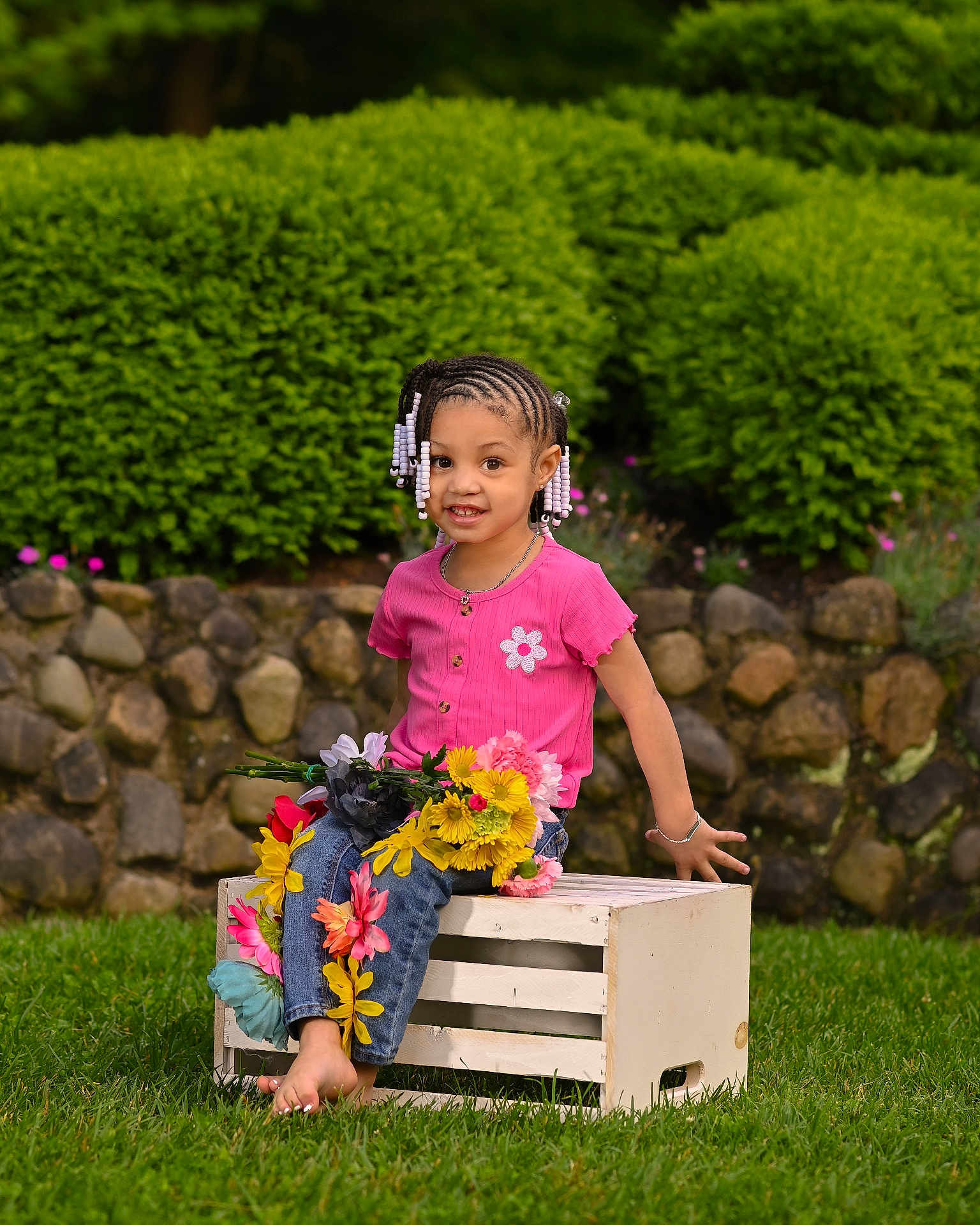 Kobi is registered to the contest to win money with this photo: child, girl, smile, pink_shirt, braided_hair, beads, flowers, jeans, barefoot, wooden_crate, grass, garden, greenery, stone_wall, outdoor, nature, happy, portrait, sitting, daylight