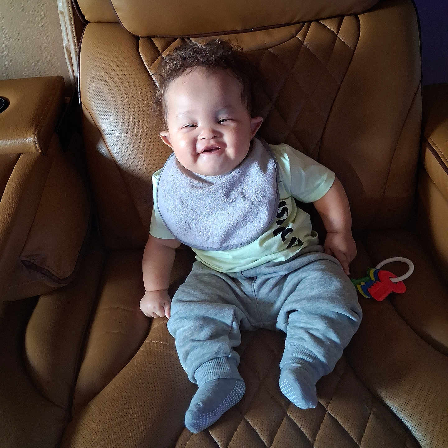 Braelin is registered to the contest to win money with this photo: baby, child, smiling, sitting, portrait, leather_chair, armchair, indoor, home, clothing, bib, pants, socks, toy, happy, face, curly_hair, furniture, cozy, photograph
