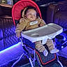 Braelin is registered to the contest to win money with this photo: baby, infant, child, high_chair, sitting, sleepy, face, portrait, indoor, living_room, furniture, red, beige, purple_lighting, blue_lighting, tray, carpet, couch, clothing, photography