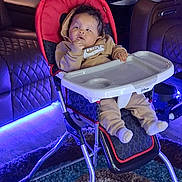 Braelin is registered to the contest to win money with this photo: baby, infant, child, high_chair, sitting, portrait, face, looking_up, hand, hoodie, clothing, white_shoes, tray, stroller, living_room, couch, carpet, indoor, blue_lighting, neon_lighting