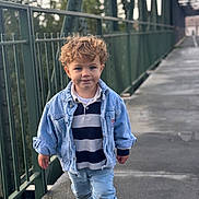 Lyam a rejoint le concours — aidez-le/la à gagner de superbes lots ! child, toddler, curly_hair, denim_jacket, striped_shirt, jeans, black_shoes, walking, bridge, metal_railings, outdoor, daylight, curious_expression, urban, portrait, person, casual_clothing, young_child, concrete_floor, background_blur