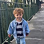 child, toddler, curly_hair, denim_jacket, striped_shirt, jeans, black_shoes, walking, bridge, metal_railings, outdoor, daylight, curious_expression, urban, portrait, person, casual_clothing, young_child, concrete_floor, background_blur