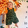 baby, blanket, camouflage, child, cute, fabric, face, feet, giraffe, green, hand, indoors, infant, laying_down, orange, orange_shirt, pattern, person, smile, soft_surface