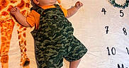 Aiden is registered to the contest to win money with this photo: baby, infant, child, giraffe, blanket, camo_overalls, orange_shirt, numbers, greenery, laying_down, cute, curious, pattern, fabric, soft, indoor, portrait, small_hands, feet, face