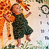 baby, blanket, camo_overalls, child, curious, cute, fabric, face, feet, giraffe, greenery, indoor, infant, laying_down, numbers, orange_shirt, pattern, portrait, small_hands, soft