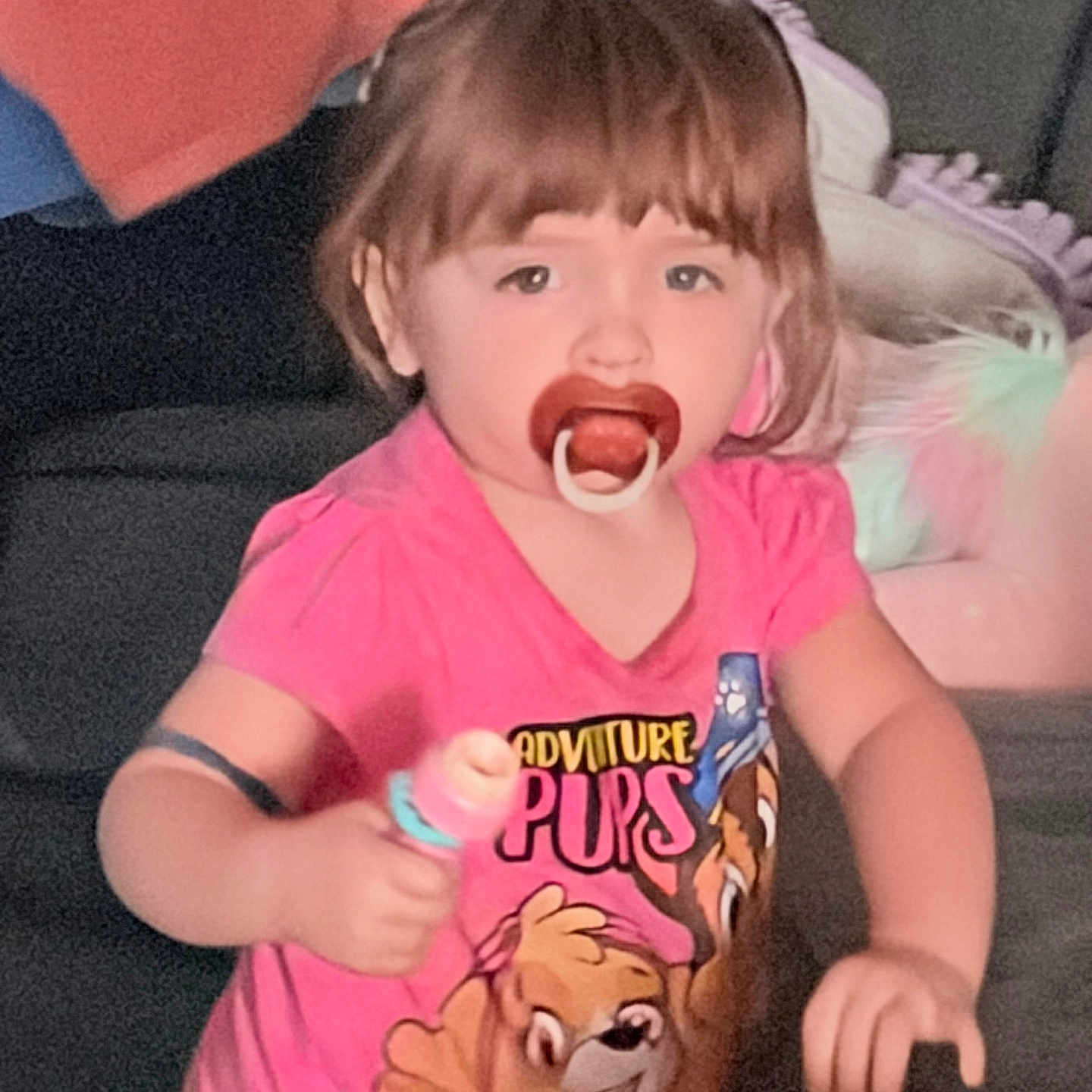 Addilynn is registered to the contest to win money with this photo: toddler, child, pacifier, pink_shirt, indoor, toy, blanket, couch, curious, person, young_child, short_hair, cartoon_print, furniture, home, casual_clothing, portrait, smiling, baby, cute