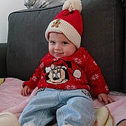 Mia a rejoint le concours — aidez-le/la à gagner de superbes lots ! baby, child, smiling, red_hat, pom_pom, red_sweater, mickey_mouse, blue_jeans, white_boots, couch, blanket, indoor, cute, happy, holiday_clothing, festive, person, seated, face, clothing