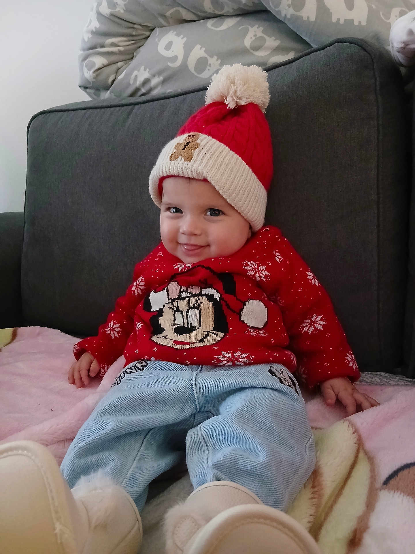 Mia a rejoint le concours — aidez-le/la à gagner de superbes lots ! baby, child, smiling, red_hat, pom_pom, red_sweater, mickey_mouse, blue_jeans, white_boots, couch, blanket, indoor, cute, happy, holiday_clothing, festive, person, seated, face, clothing