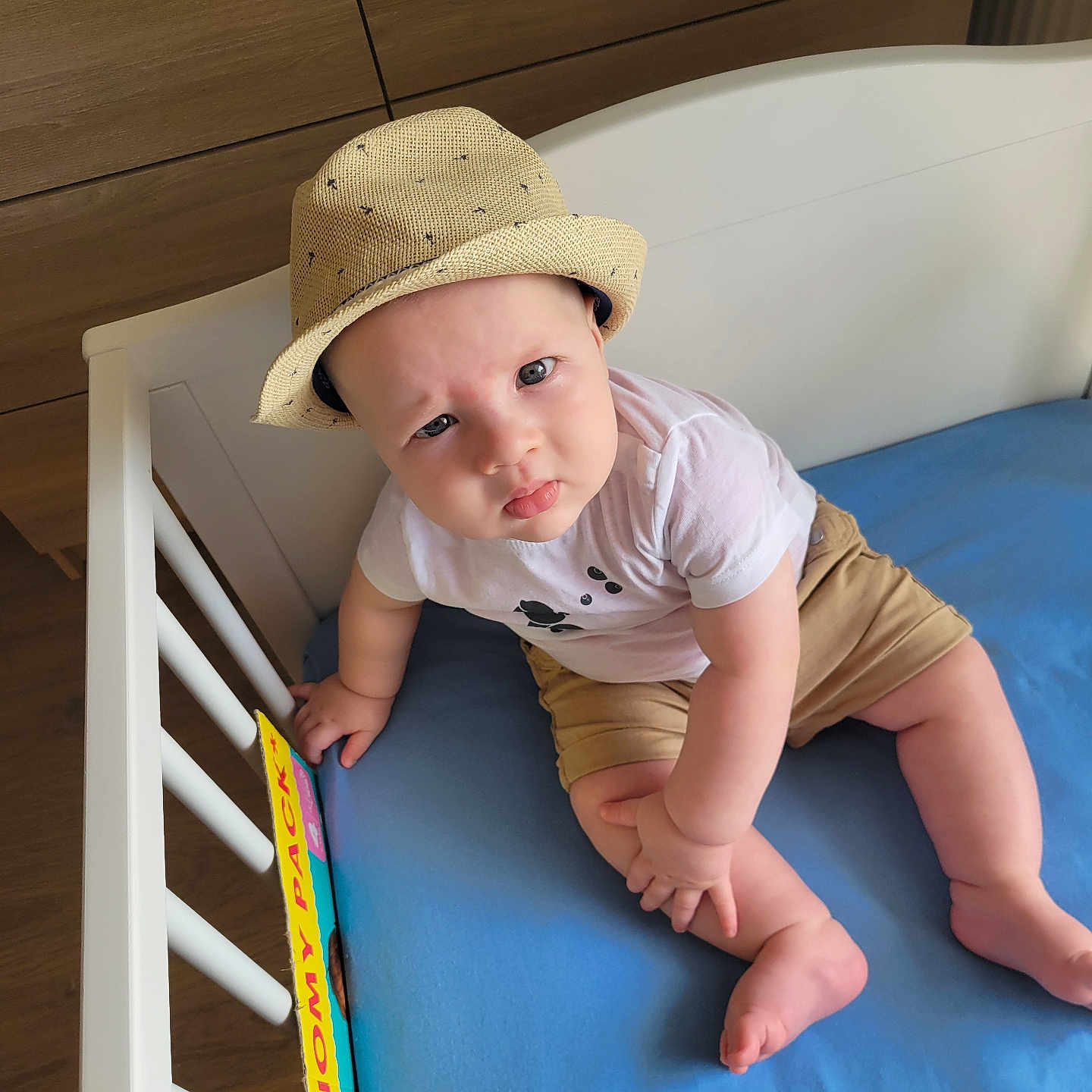 Aylan a rejoint le concours — aidez-le/la à gagner de superbes lots ! baby, barefoot, blue_mattress, child, crib, curious_expression, floor, furniture, hat, headwear, indoor, infant, light_clothing, person, puzzle_expression, room, sitting, toy, white_crib, wooden_background