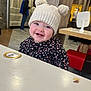 baby, background, casual_clothing, child, cute, face, floor, happy, hat, indoor, knit_hat, people, person, pom_poms, portrait, restaurant, seated, smile, table, wood_paneling