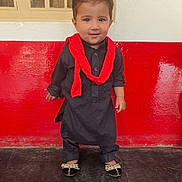 Yusuf joined the competition — help win amazing prizes! toddler, child, traditional_clothing, black_outfit, red_scarf, standing, smiling, indoor, wall, red_wall, white_wall, footwear, decorative_sandals, hair, portrait, person, floor, cute, young_child, happy