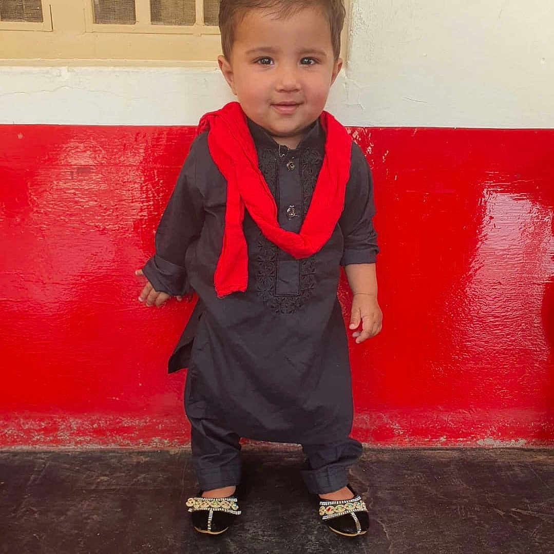 Yusuf joined the competition — help win amazing prizes! black_outfit, child, cute, decorative_sandals, floor, footwear, hair, happy, indoor, person, portrait, red_scarf, red_wall, smiling, standing, toddler, traditional_clothing, wall, white_wall, young_child