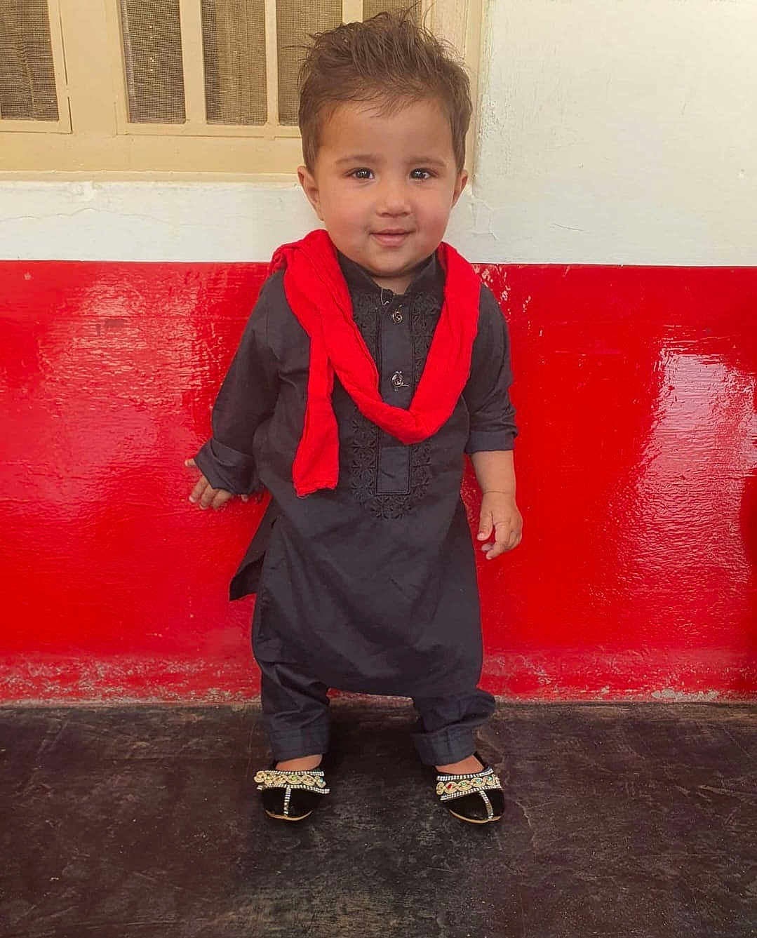 Yusuf joined the competition — help win amazing prizes! toddler, child, traditional_clothing, black_outfit, red_scarf, standing, smiling, indoor, wall, red_wall, white_wall, footwear, decorative_sandals, hair, portrait, person, floor, cute, young_child, happy
