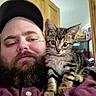 animal, beard, brown_hair, cap, casual_clothing, closeup, companion, cute, door, facial_hair, furniture, indoor, kitten, man, pet, portrait, relaxed, shelf, tabby_cat, young_cat