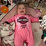 baby, infant, smiling, pink_onesie, harley_davidson, crib, blanket, plush_toy, stuffed_animal, happy, child, cute, indoor, person, soft_toys, face, lying_down, cozy, baby_clothing, playful