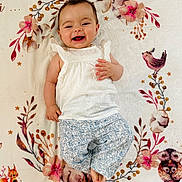 Kélia participe au concours pour gagner de l'argent avec cette photo : baby, smile, child, blanket, floral_pattern, woodland_animals, cute, lying_down, happy, white_clothing, patterned_pants, infant, face, portrait, indoors, soft_texture, decorative, cozy, small_child, playful