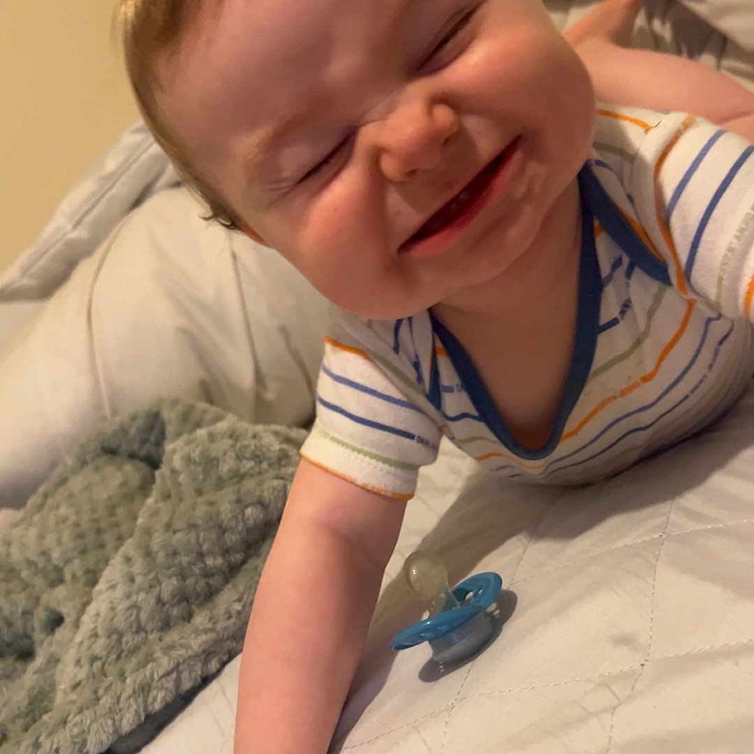 Noah joined the competition — help win amazing prizes! arm, baby, bed, blanket, cheeks, clothing, cute, expression, face, hand, happy, indoors, infant, lying_down, pacifier, person, skin, smiling, striped_shirt, toddler