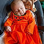 baby, sleeping, orange_dress, pumpkin_outfit, infant, cushion, blanket, knitted_toy, baby_seat, cozy, cute, peaceful, child, portrait, indoors, soft_lighting, clothing, hand, face, toddler