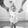 baby, child, toddler, crown, birthday, standing, walking, supported, smiling, portrait, black_and_white, indoor, nursery, adult_hands, parents, clothing, skirt, wooden_floor, fluffy_rug, photography
