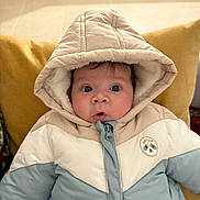 Joys a rejoint le concours — aidez-le/la à gagner de superbes lots ! baby, child, hood, jacket, puffy_jacket, coat, indoor, face, wide_eyes, curious, seated, pillow, mustard_color, warm_clothing, winter_wear, zipper, cute, portrait, infant, cozy