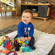 Gabriel a rejoint le concours — aidez-le/la à gagner de superbes lots ! baby, child, smiling, toys, book, blanket, floor, chair, box, cabinet, television, wooden_floor, carpet, indoor, happy, person, clothing, blue_sweater, play, cute