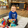 baby, child, smiling, toys, book, blanket, floor, chair, box, cabinet, television, wooden_floor, carpet, indoor, happy, person, clothing, blue_sweater, play, cute