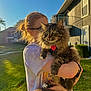 Socks is registered to the contest to win money with this photo: cat, person, glasses, outdoor, sunlight, grass, house, fluffy_cat, tabby_cat, holding, pet, daylight, leash, collar, green_eyes, casual_clothing, backyard, sunset, happy, nature
