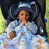 toddler, child, baby, hat, bucket_hat, stroller, blanket, thumb_sucking, cozy_clothing, outdoor, portrait, face, cute, young_child, seat, baby_blanket, nature, greenery, close_up, casual